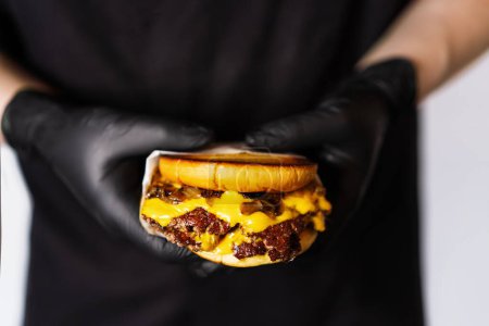 Foto de Una jugosa hamburguesa artesanal con queso se lleva a cabo en manos de un chef con guantes negros. - Imagen libre de derechos