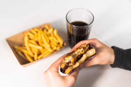 Foto de El tipo tiene una hamburguesa en la mano, cola y papas fritas en la mesa. - Imagen libre de derechos