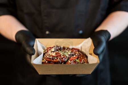 Photo for The cook holds in his hands a box with grilled spicy chicken teriyaki legs in packs for delivery. - Royalty Free Image