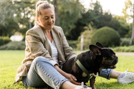 Foto de Chica rubia de cuarenta años se acurruca con su perro, un bulldog francés, en un parque sobre la hierba. - Imagen libre de derechos