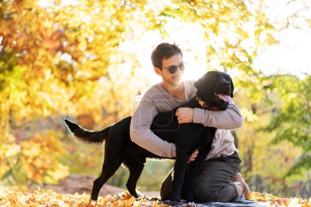 Téléchargez les photos : Guy aux lunettes de soleil étreint son chien dans le parc d'automne au coucher du soleil - en image libre de droit