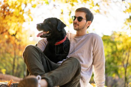 Téléchargez les photos : Guy aux lunettes de soleil étreint son chien dans le parc d'automne au coucher du soleil - en image libre de droit