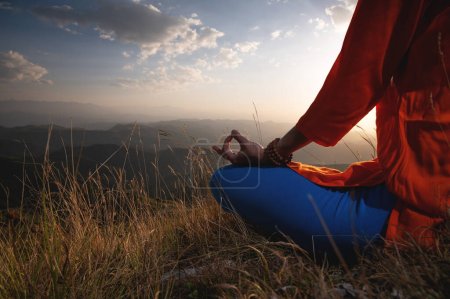 Foto de Close-up of a woman and female hands sitting in a lotus position, yoga outdoors in the grass at sunset against the backdrop of a mountain range and clouds, meditation. - Imagen libre de derechos