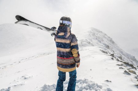 Foto de El esquiador se encuentra en la cima de la montaña y disfruta de la vista de las hermosas montañas de invierno en un día nublado. Mujer con traje de esquí sosteniendo esquís en su hombro. - Imagen libre de derechos