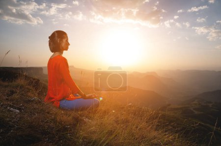 Téléchargez les photos : Une femme pratique le yoga et médite sur une montagne. Vue latérale d'un yogi pratiquant dans les montagnes à l'aube dans les rayons du soleil recevant l'énergie d'un nouveau jour. - en image libre de droit