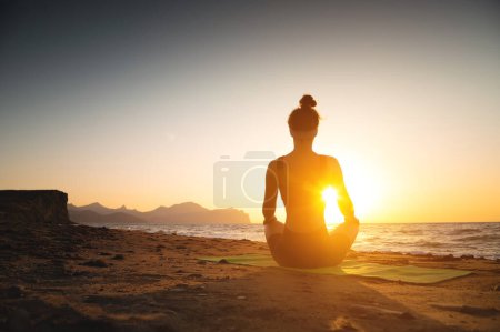 Téléchargez les photos : Une femme médite sur la côte au bord de la mer, vue arrière. Endroit pour le texte. Yoga, harmonie de la santé, silhouette d'une jeune femme sur la plage au coucher du soleil ou à l'aube. - en image libre de droit