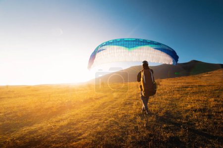Photo for A male paraglider takes off from a yellow field with a blue parachute against the backdrop of hills and small mountains. Paragliding - Royalty Free Image