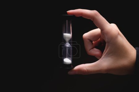Photo for Close-up of a woman holding an hourglass in the dark. Fingers hold on both sides a plastic sand clock with white sand on a black background. - Royalty Free Image
