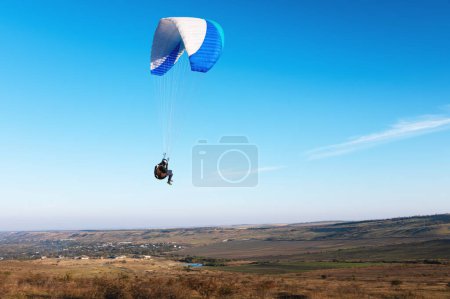 A paraglider takes off from a mountainside with a blue and white canopy and the sun behind. A paraglider is a silhouette. The glider is sharp, with little wing movement. A male paraglider launches a
