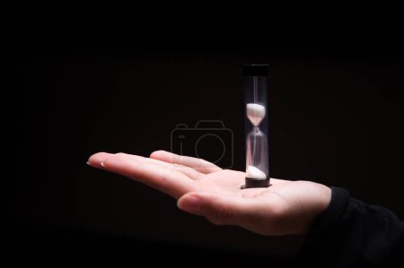 Close-up of a woman holding an hourglass in the dark. Fingers hold on both sides a plastic sand clock with white sand on a black background.