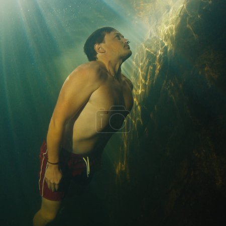 Téléchargez les photos : L'homme plonge dans la rivière d'eau douce avec des rayons ensoleillés qui brillent dans l'eau et nage près de la roche - en image libre de droit
