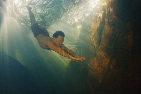 Téléchargez les photos : L'homme plonge dans la rivière d'eau douce avec des rayons ensoleillés qui brillent dans l'eau et nage près de la roche - en image libre de droit