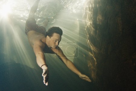 Foto de El hombre se sumerge en el río de agua dulce con rayos soleados brillando a través del agua y nada cerca de la roca - Imagen libre de derechos