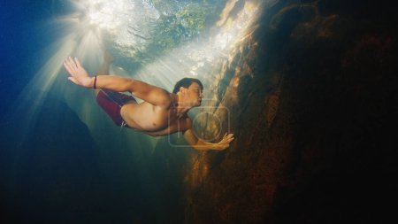 Photo for Man dives in the freshwater river with sunny rays shining through the water and swims near the rock - Royalty Free Image