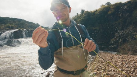 Foto de Pescador se prepara para la pesca con mosca. Pescador en vadeadores se encuentra junto al río rápido y oscuro y prepara el aparejo para la pesca - Imagen libre de derechos