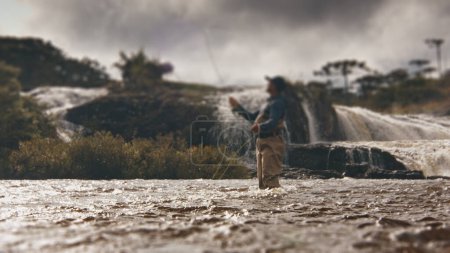 Foto de Pesca con mosca. Pescador en vadeadores lanza la línea y la pesca en el río turbio rápido con cascada en el fondo. Concéntrate en el agua en primer plano - Imagen libre de derechos