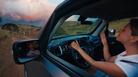 Téléchargez les photos : Femme dirige la voiture sur une route rurale au coucher du soleil vif. Une voyageuse seule conduit la voiture sur une route non asphaltée vide - en image libre de droit