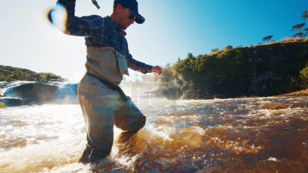 Photo for Fly fishing in a rapid river. Fisherman in waders slowly walks from the furious murky river - Royalty Free Image