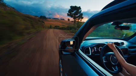 Téléchargez les photos : Femme dirige la voiture sur une route rurale au coucher du soleil vif. Une voyageuse seule conduit la voiture sur une route non asphaltée vide - en image libre de droit