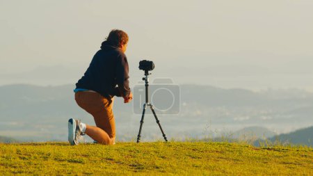 Foto de Fotógrafo en la colina. Joven fotógrafo masculino toma fotos con cámara y teleobjetivo en trípode en la colina verde en las montañas al amanecer - Imagen libre de derechos