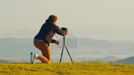 Téléchargez les photos : Photographe sur la colline. Jeune photographe prend des photos avec appareil photo et téléobjectif sur trépied sur la colline verdoyante dans les montagnes au lever du soleil - en image libre de droit