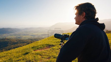Foto de Fotógrafo en la colina. Joven fotógrafo masculino toma fotos con cámara y teleobjetivo en trípode en la colina verde en las montañas al amanecer - Imagen libre de derechos
