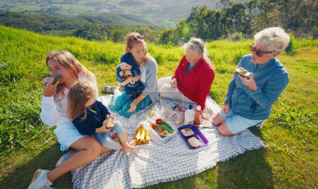 Téléchargez les photos : Grande famille pique-niquer sur la colline verdoyante dans les montagnes au lever du soleil - en image libre de droit