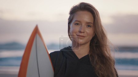 Téléchargez les photos : Femme surfeuse se tient debout avec planche de surf sur la plage tropicale dans poncho et regarde dans la caméra - en image libre de droit