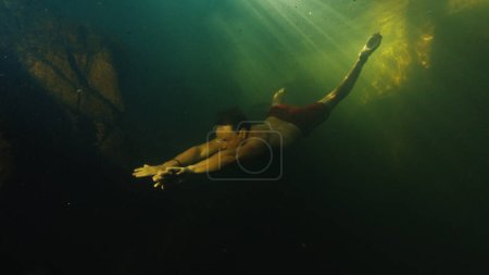 Téléchargez les photos : Jeune homme plongée libre dans le lac situé dans une forêt et nage par des roches sous-marines - en image libre de droit