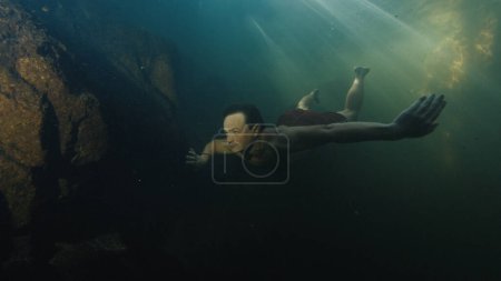 Téléchargez les photos : Jeune homme plongée libre dans le lac situé dans une forêt et nage par des roches sous-marines - en image libre de droit