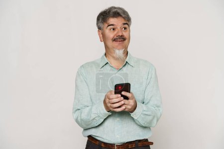 Mature man with grey beard smiling and using mobile phone isolated over white background