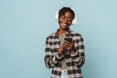 African american young woman in plaid shirt using headphones and cellphones isolated over blue background