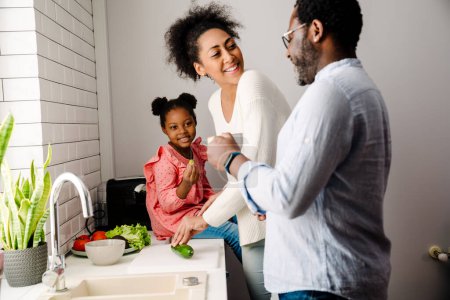 Téléchargez les photos : Africain américain heureux homme et femme avec petite fille faire le petit déjeuner dans la cuisine à la maison - en image libre de droit