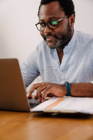 Téléchargez les photos : Homme barbu afro-américain travaillant sur ordinateur portable tout en étant assis par bureau à la maison - en image libre de droit