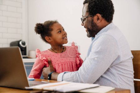 Téléchargez les photos : Homme afro-américain étreignant sa petite fille tout en travaillant sur ordinateur portable à la maison - en image libre de droit
