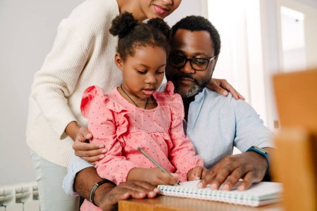Téléchargez les photos : Petite fille afro-américaine écrivant dans un bloc-notes avec stylo tout en étant assis avec les parents à table à la maison - en image libre de droit