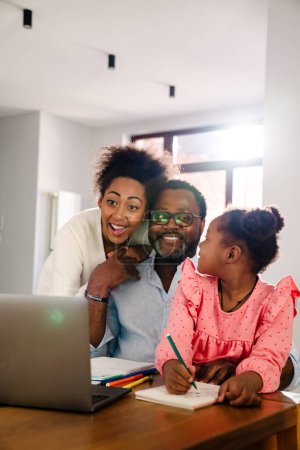 Photo for African american family with little girl smiling and using laptop while sitting by table at home - Royalty Free Image