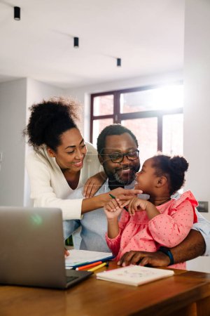 Téléchargez les photos : Famille afro-américaine avec une petite fille souriante et utilisant un ordinateur portable assis à table à la maison - en image libre de droit