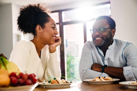 Téléchargez les photos : Femme de famille afro-américaine et homme souriant et assis à table tout en prenant le petit déjeuner à la maison - en image libre de droit