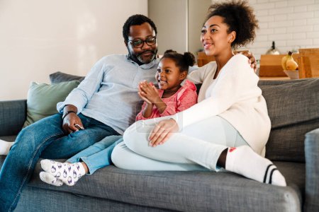 Foto de Afroamericana familia mujer y hombre con pequeña hija sonriendo y sentado en sofá en casa - Imagen libre de derechos