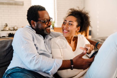 Téléchargez les photos : Femme adulte afro-américaine et homme souriant et utilisant un téléphone portable assis sur le canapé à la maison - en image libre de droit