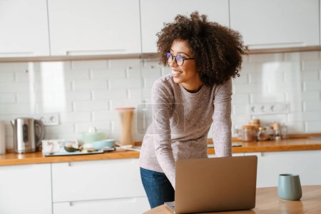 Téléchargez les photos : Africaine américaine jeune femme avec frisée afro coiffure et lunettes à l'aide d'un ordinateur portable à la maison - en image libre de droit