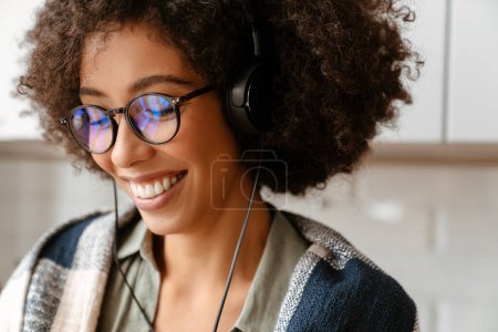 Téléchargez les photos : Africaine américaine jeune femme avec frisée afro coiffure et lunettes à l'aide d'écouteurs à la maison - en image libre de droit