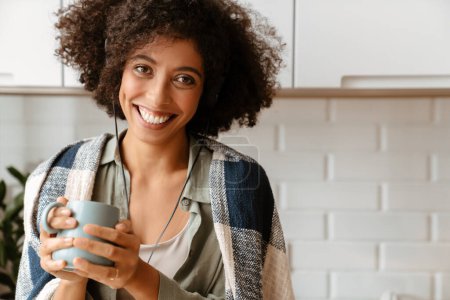 Foto de Mujer joven afroamericana con peinado afro rizado usando auriculares mientras bebe café en casa - Imagen libre de derechos