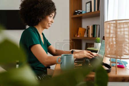 African american young woman with curly afro hairstyle sitting at desk and using laptop at home Poster 624874762