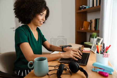 Téléchargez les photos : Afro-américaine jeune femme avec frisée afro coiffure assis au bureau et en utilisant un ordinateur portable à la maison - en image libre de droit