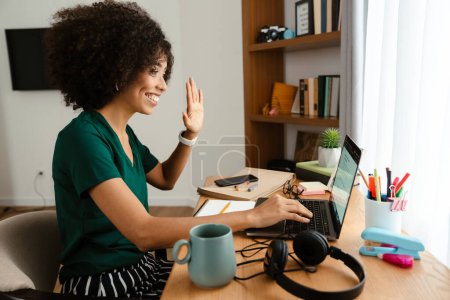Photo for African american young woman with curly afro hairstyle sitting at desk and using laptop at home - Royalty Free Image