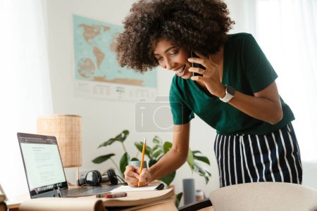 Téléchargez les photos : Africaine américaine jeune femme avec frisée afro coiffure parler sur téléphone portable et prendre des notes à la maison - en image libre de droit