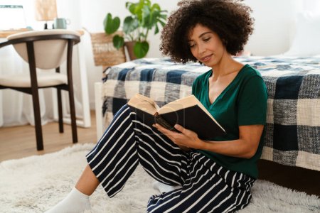 Foto de Mujer joven afroamericana con peinado afro rizado sonriendo y leyendo libro en casa - Imagen libre de derechos