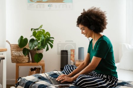 Téléchargez les photos : Afro-américaine jeune femme avec frisée afro coiffure assis dans le lit et en utilisant un ordinateur portable à la maison - en image libre de droit